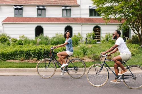 A man and woman ride bicycles on a sunny street, smiling, with a house and greenery in the background.