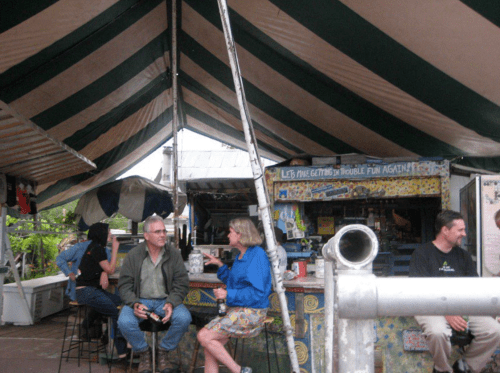A lively outdoor market scene under a striped tent, with people chatting and a colorful bar in the background.