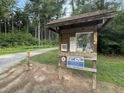 A wooden trail information sign with a map, located near a gravel path surrounded by trees.
