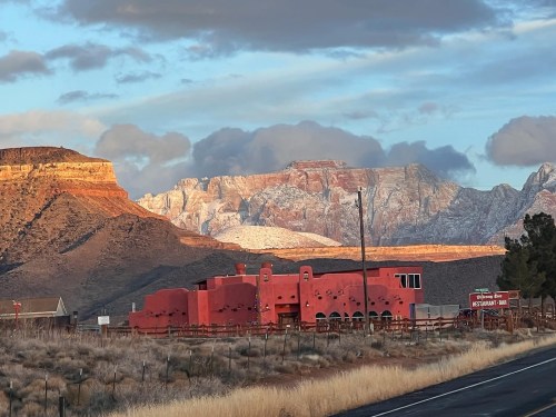 A vibrant pink building near a road, set against dramatic mountains and a cloudy sky at sunset.