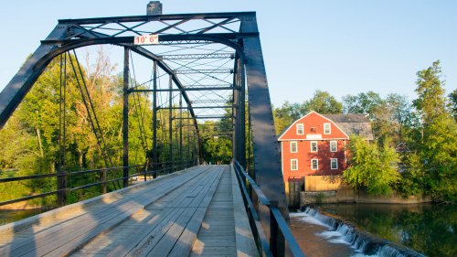 A black metal bridge spans a river, with a red building and lush greenery in the background under a clear blue sky.