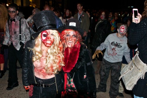 Two people in zombie costumes with dramatic makeup and blood effects pose for the camera, surrounded by a crowd.