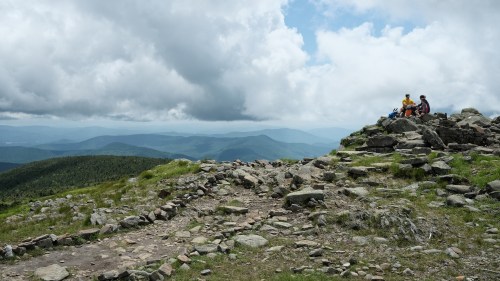 A group of hikers sits on rocky terrain, overlooking a lush green valley and distant mountains under a cloudy sky.