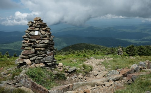 A stone cairn stands on a mountain trail, overlooking a lush green valley and distant hills under a cloudy sky.