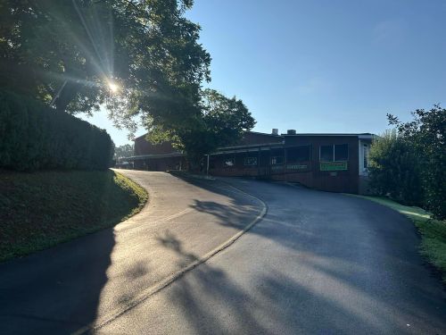 A winding road leads to a wooden building with a restaurant sign, surrounded by trees and bathed in morning light.
