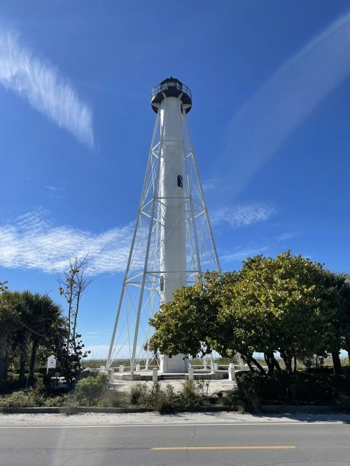 A tall white lighthouse stands against a clear blue sky, surrounded by greenery and palm trees.