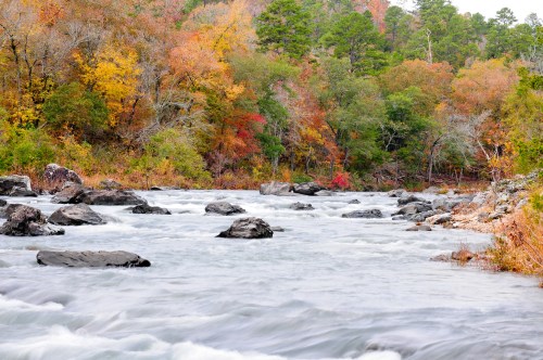A serene river flows over rocks, surrounded by vibrant autumn foliage in shades of orange, yellow, and green.