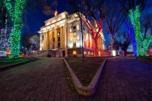 A beautifully lit historic building at night, surrounded by colorful holiday lights on trees.