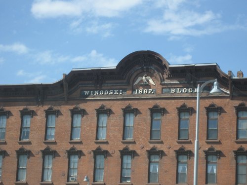 Historic brick building with "Winooski 1867" and "Block" inscribed, under a blue sky with scattered clouds.