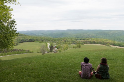 Two people sit on a grassy hill, overlooking a lush green valley and distant mountains under a cloudy sky.