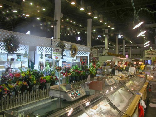 A bustling market interior with flower displays, food counters, and bright overhead lights.