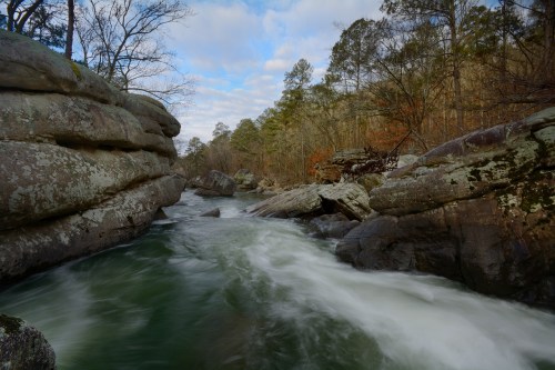 A serene river flows between rocky banks, surrounded by trees under a partly cloudy sky.