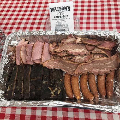 A tray of assorted barbecue meats, including ribs, brisket, and sausages, on a red and white checkered tablecloth.