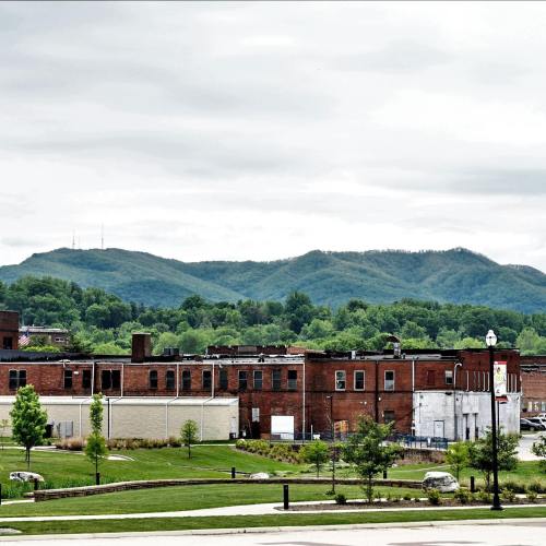 A view of a small town with brick buildings in the foreground and green mountains in the background under a cloudy sky.