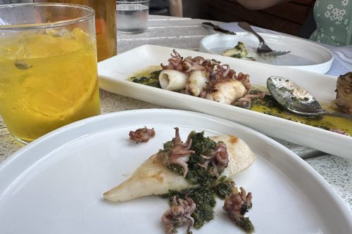 A plate of squid with greens, garnished with small octopus, alongside a drink in a casual dining setting.