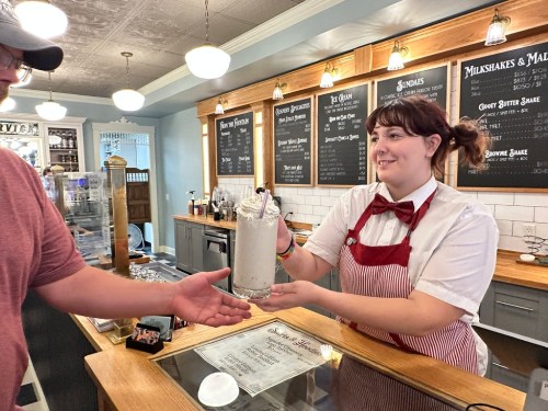 A server in a retro diner outfit hands a milkshake to a customer at the counter. Menus are displayed on the wall.