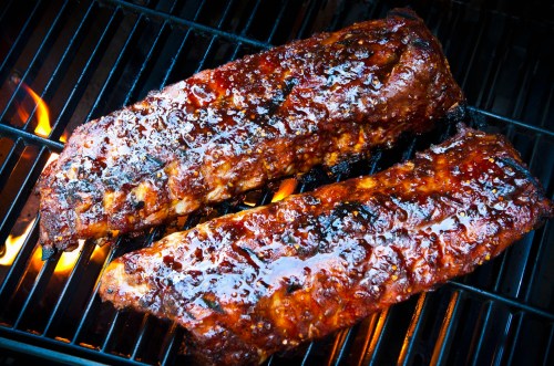 Two glazed, grilled ribs sizzling on a barbecue grill, with flames visible underneath.