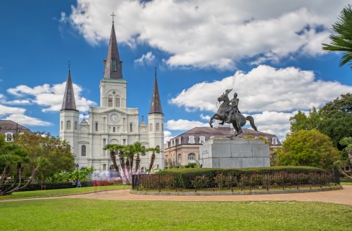 Jackson Square features St. Louis Cathedral and a statue of Andrew Jackson against a cloudy sky. Lush greenery surrounds the area.
