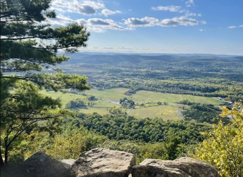 A scenic view of rolling hills and a valley under a blue sky with scattered clouds, framed by trees and rocks.