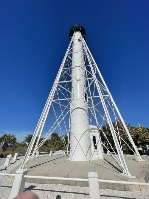 A tall, white lighthouse with a metal framework stands against a clear blue sky, surrounded by greenery.