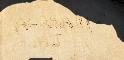 The sand features the words "ALOHA!!!" and "MJ" written in a playful, casual style.
