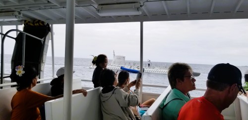Passengers on a boat view a floating platform in the ocean, with cloudy skies overhead.