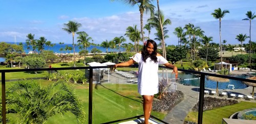 A woman stands on a balcony overlooking a tropical resort with palm trees and a pool, enjoying a sunny day.