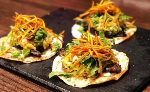 Three tacos on a black slate, topped with shredded lettuce, carrots, and a mix of colorful vegetables.