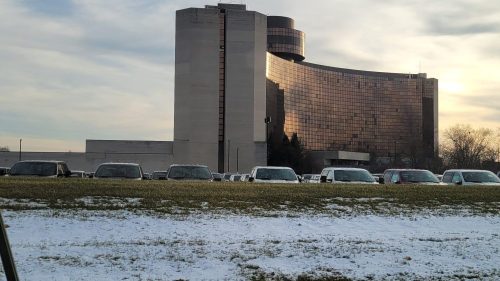 A modern building with a reflective facade, surrounded by parked cars and a snowy grassy area in the foreground.