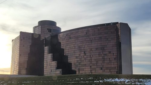 A modern, angular building with reflective glass windows, set against a cloudy sky and grassy foreground.