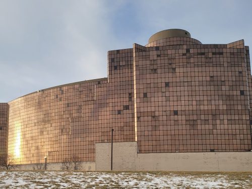 A large, modern building with a reflective, copper-colored facade and a curved roof, set against a cloudy sky.