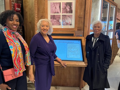 Three women stand in front of a display featuring historical photos and information, smiling and posing together.