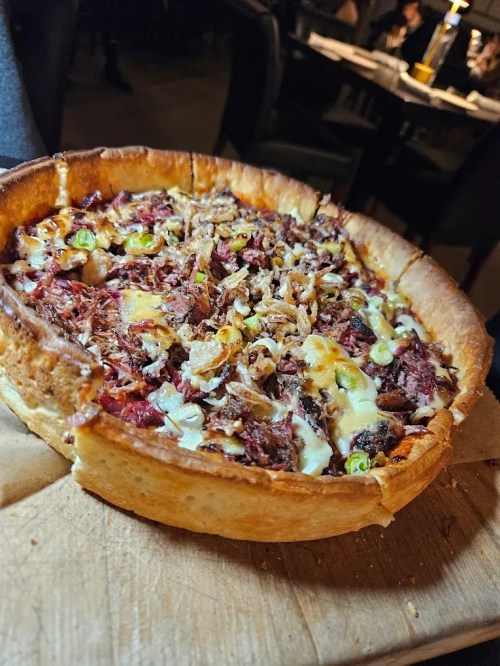 A deep-dish pizza topped with cheese, pulled meat, and green onions, served on a wooden board.