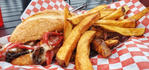 A sandwich with meat, cheese, and toppings, served with a side of golden-brown fries in a red and white checkered basket.