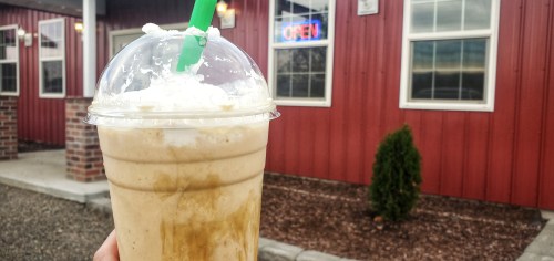 A hand holding a large iced coffee drink topped with whipped cream, in front of a red building with an "Open" sign.