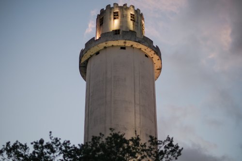 A tall, illuminated lighthouse against a twilight sky, surrounded by trees.