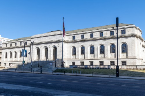 A large, historic stone building with steps, columns, and a flag, set against a clear blue sky.