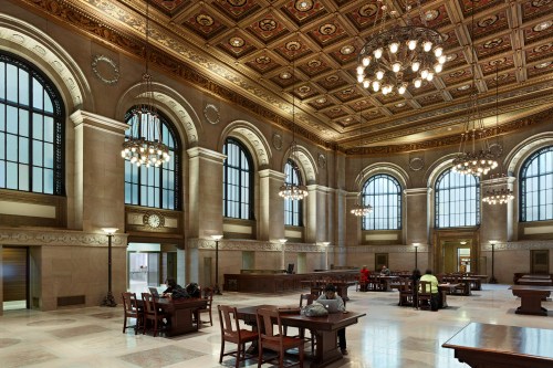 A spacious, elegantly designed interior of a historic building with large windows, ornate ceilings, and wooden tables.
