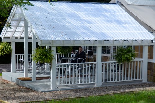 A person sits on a covered patio surrounded by ferns and outdoor furniture, reading a newspaper.