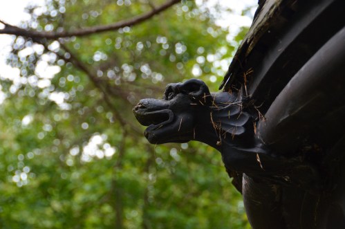 A carved dragon head on a roof, partially covered in pine needles, with a blurred green background.