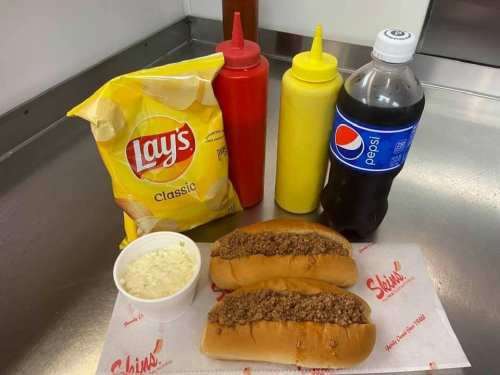Two hot dogs with meat sauce, a cup of coleslaw, a bag of Lay's chips, and bottles of mustard and ketchup next to a Pepsi.
