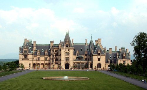 A grand mansion with intricate architecture, surrounded by lush green lawns and a fountain in the foreground.