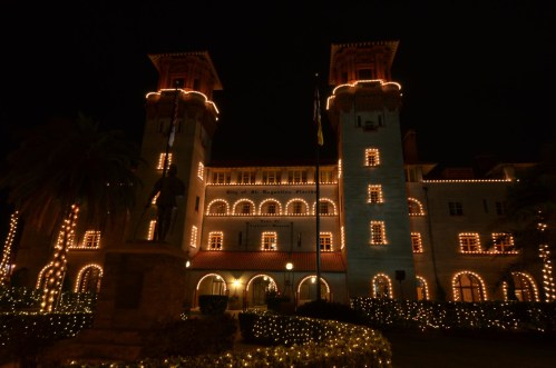 Historic building illuminated with festive lights at night, surrounded by greenery and a statue in the foreground.