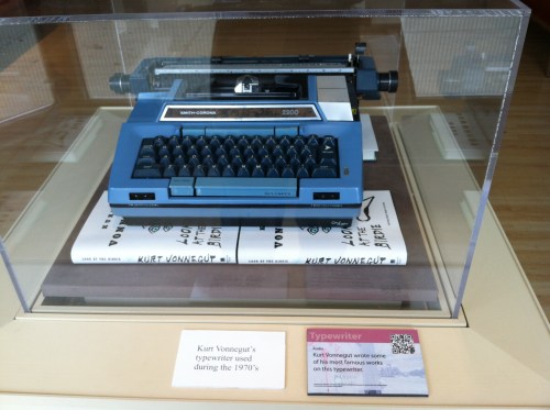 A blue Smith-Corona typewriter displayed in a glass case, with papers featuring Kurt Vonnegut's name underneath.