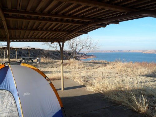 A tent under a shelter with a view of a lake and dry grassland in the background. Clear blue sky above.