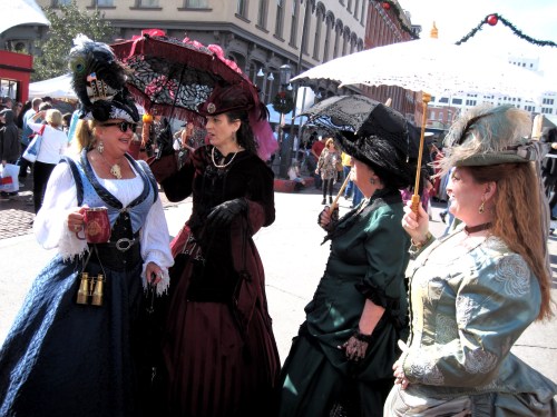 Four women in Victorian-style dresses and parasols chat at a lively outdoor event.