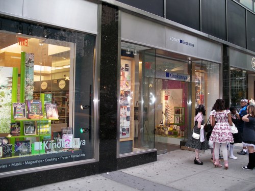 A bookstore exterior featuring large windows displaying books and magazines, with people walking by on the sidewalk.