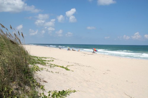 A sandy beach with gentle waves, colorful umbrellas, and a clear blue sky dotted with clouds.
