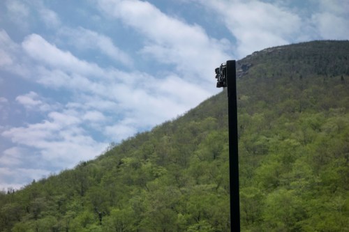 A tall pole with lights stands against a backdrop of a green mountain and a partly cloudy sky.