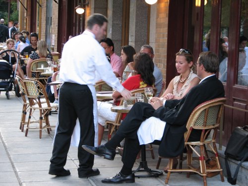 A waiter serves drinks to patrons at an outdoor café, with people dining and socializing in a lively setting.